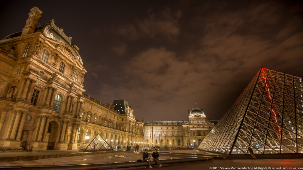 Oblique nighttime view of the Louvre Pyramid beside ornate Louvre Palace façades, with a smaller pyramid and visitors in the courtyard.