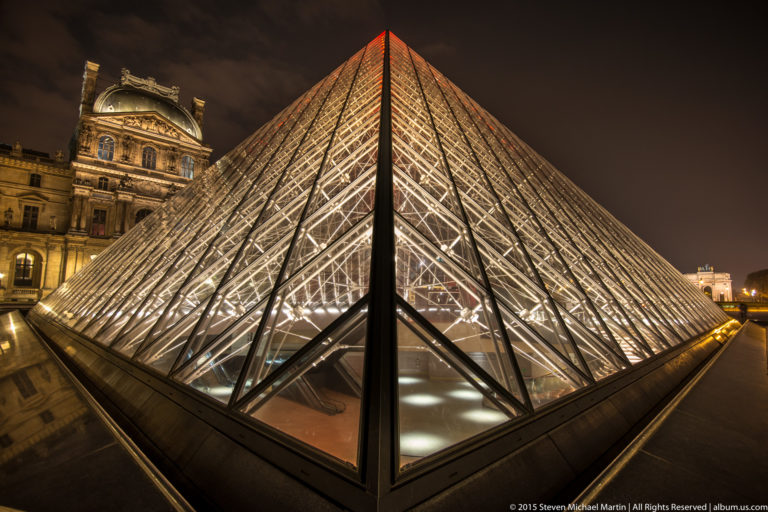 Close nighttime view of a Louvre Pyramid corner, with illuminated glass lattice converging toward the foreground and palace architecture behind.
