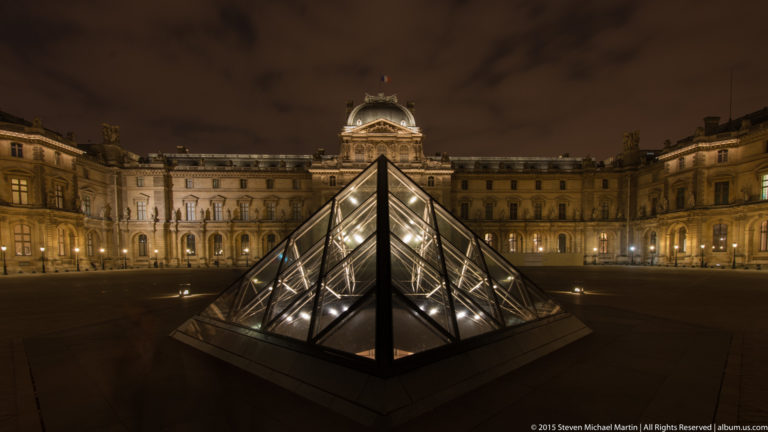 Small illuminated glass pyramid centered in the Louvre courtyard at night, with ornate palace façades filling the background.