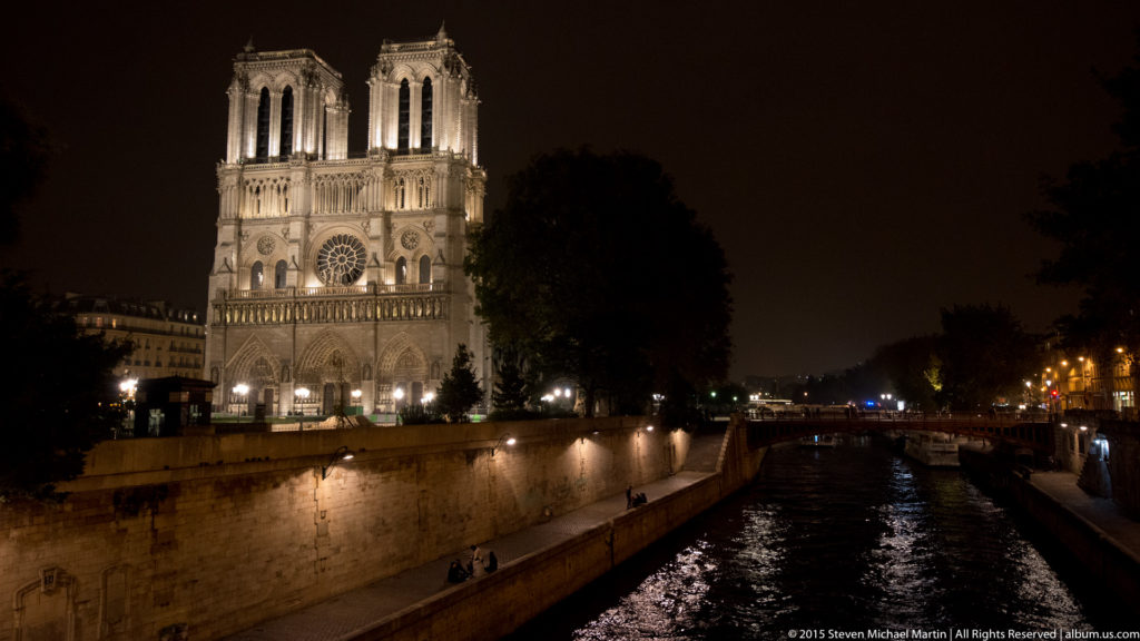 Illuminated west façade and twin towers of Notre-Dame de Paris above the Seine at night, with a stone embankment in the foreground.