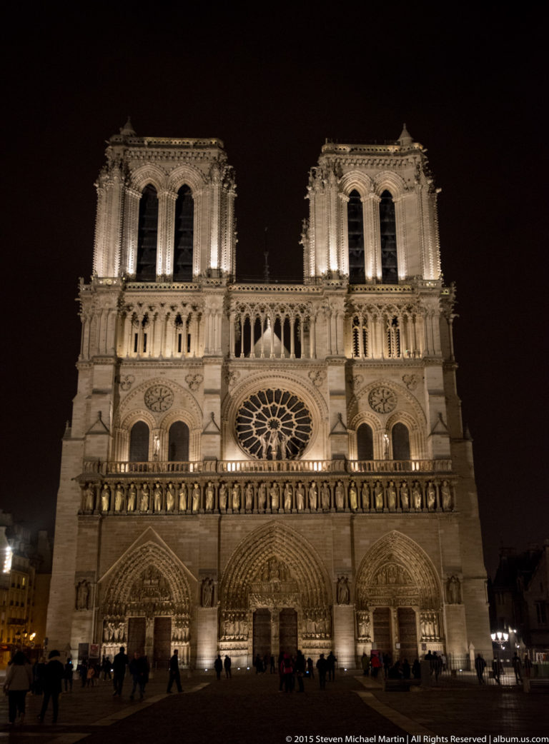 Front nighttime view of Notre-Dame de Paris, showing its illuminated twin towers, central rose window, galleries, and three Gothic portals.