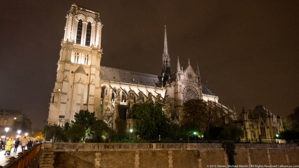 Oblique nighttime view of Notre-Dame de Paris across the Seine, showing a western tower, long Gothic flank, eastern end, and spire.