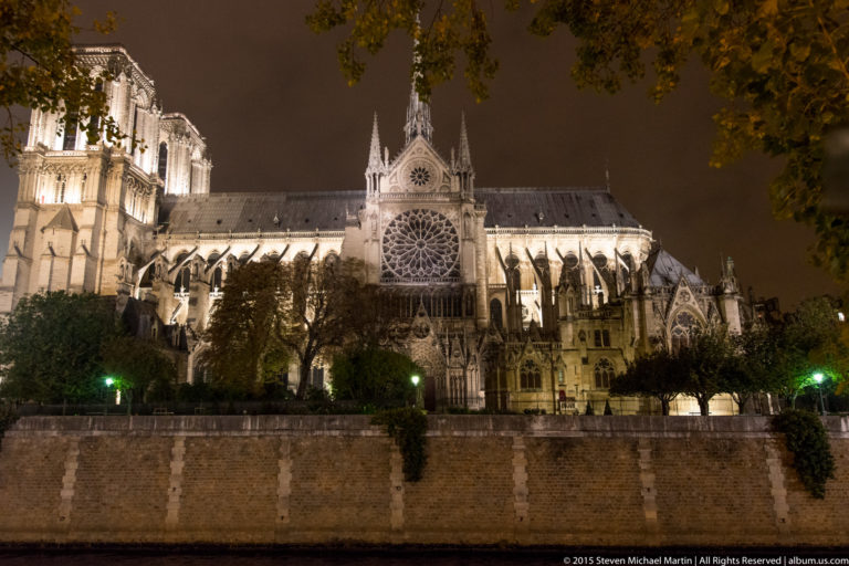 Notre-Dame de Paris at night from across the Seine, showing the transept rose window, flying buttresses, pinnacles, and former spire.
