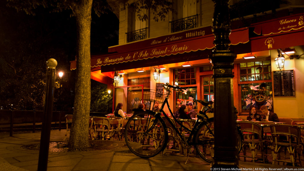 Warmly lit Paris café terrace at night with red awnings, seated patrons, outdoor tables, bicycles, and a tree in the foreground.