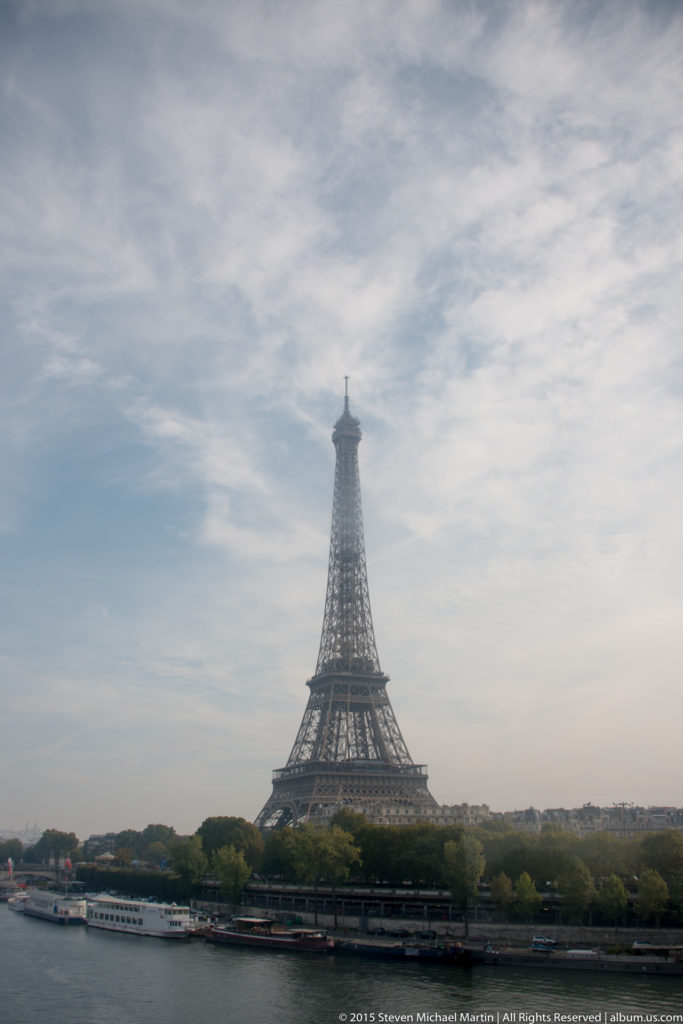Distant Eiffel Tower beyond the Seine, with riverboats, embankments, trees, and a pale clouded sky.