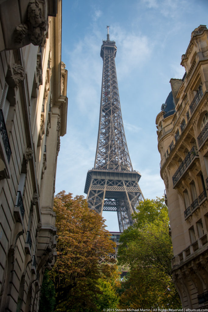 Eiffel Tower seen from a Paris street, framed by apartment buildings, trees, and blue sky.