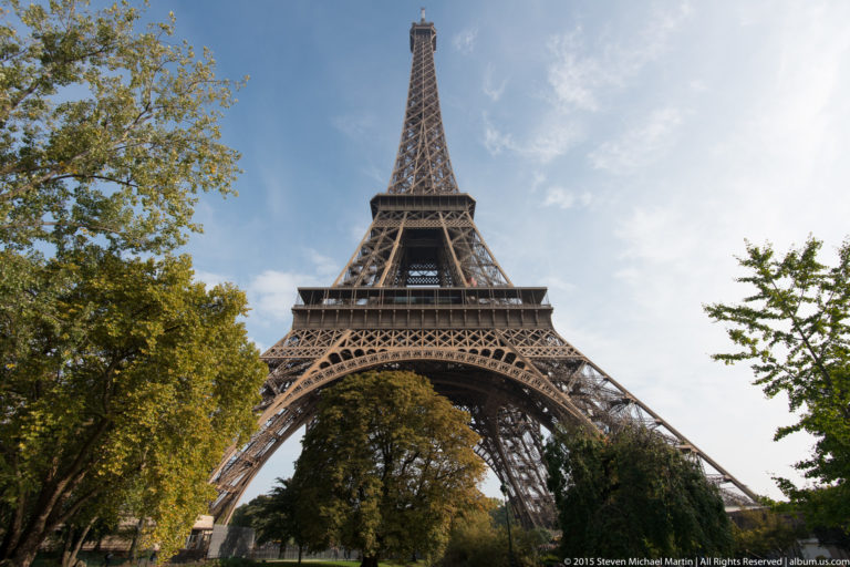 Low frontal view of the Eiffel Tower centered between trees, showing the lower arch and upper lattice structure.