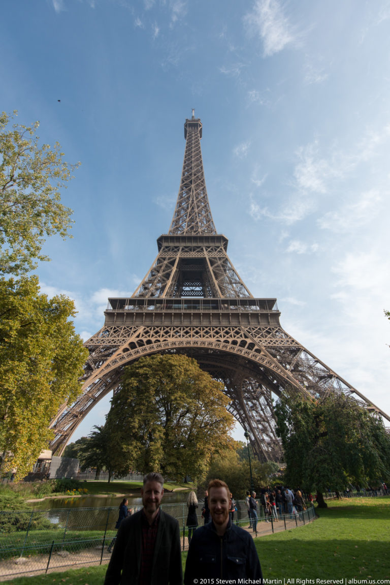 Two visitors stand before the Eiffel Tower, whose four legs spread across a tree-lined lawn under blue sky.