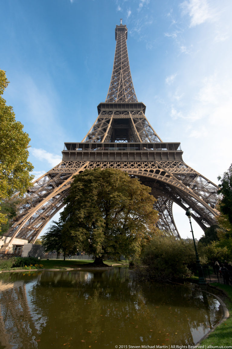 Eiffel Tower above a landscaped pond, with trees and shrubs beneath the broad iron arch.
