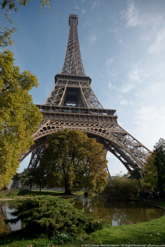 Centered Eiffel Tower beyond a garden pond and dense shrubs, with its broad iron base spanning the frame.