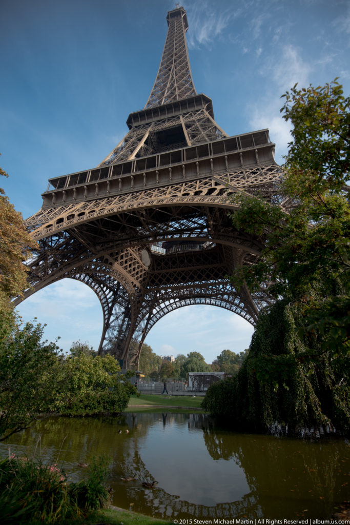 Oblique Eiffel Tower view above a pond and shrubs, with one massive iron leg dominating the left side.