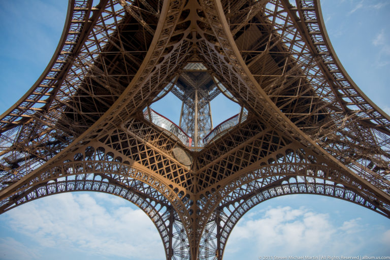 Upward view beneath the Eiffel Tower, where four iron legs and arches frame a central opening of sky.