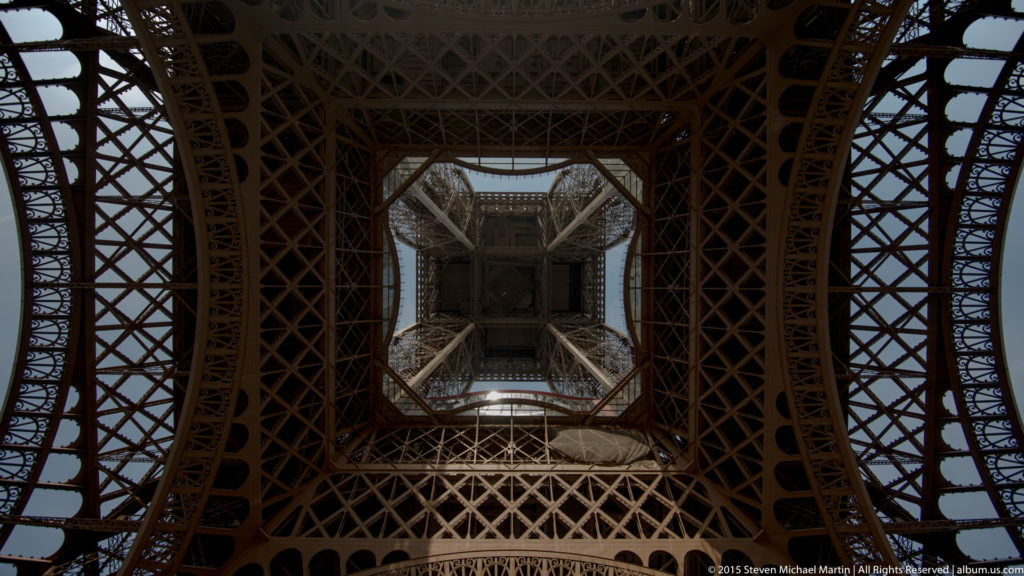 Vertical view into dense Eiffel Tower latticework, with a square central platform framed by iron beams and braces.