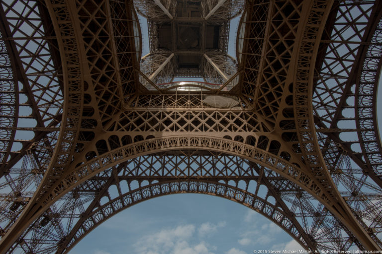 Upward Eiffel Tower view with two broad iron arches, dense lattice bracing, and blue sky between the supports.