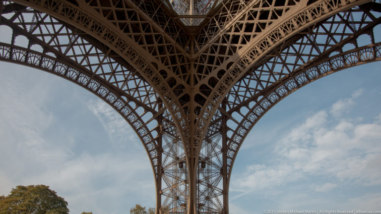 Close upward view of an Eiffel Tower arch meeting a vertical iron pier, with lattice bracing against the sky.
