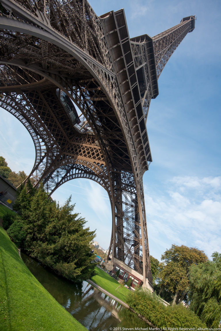 Steep oblique view up an Eiffel Tower leg above lawn, shrubs, and water, with the structure receding into blue sky.