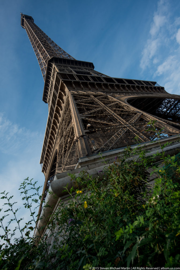 Extreme low-angle view along one Eiffel Tower pier, with green foliage at the base and blue sky beyond.