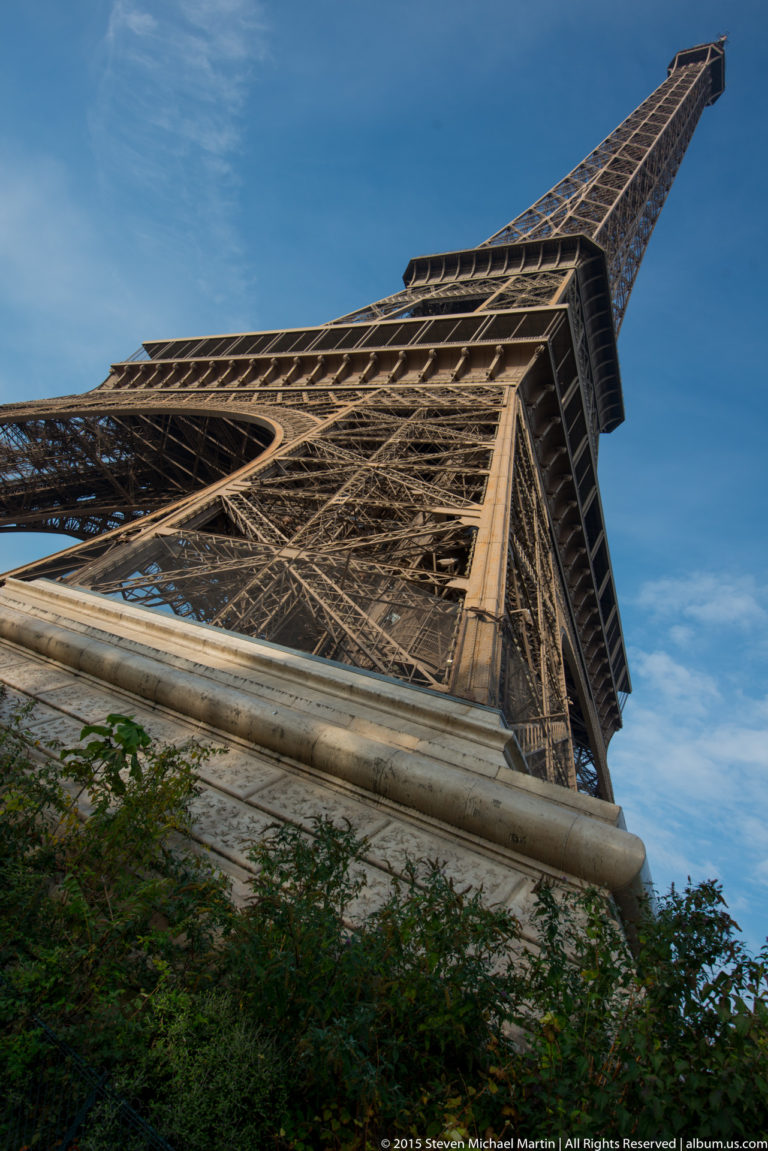 Low-angle Eiffel Tower leg rising from a stone base and shrubs toward the platform and upper lattice structure.