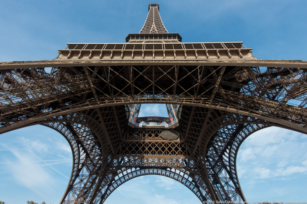 Centered upward view of the Eiffel Tower’s first platform and broad lower arch, with dense iron lattice visible beneath.