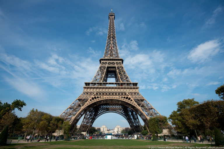 Full frontal Eiffel Tower across a lawn, framed by trees with distant city buildings visible beneath the central arch.