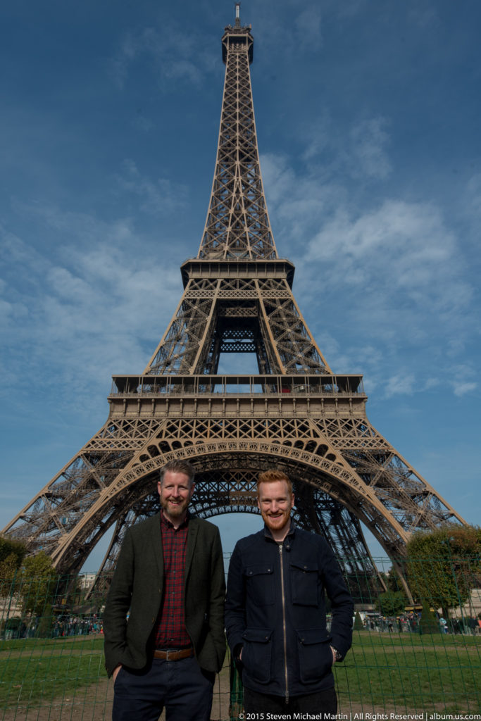 Two visitors stand side by side before the centered Eiffel Tower, with lawn and trees at the base.