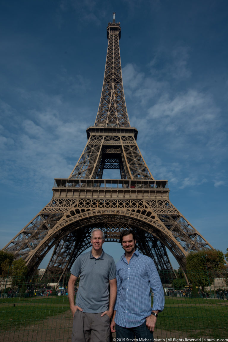 Two visitors pose in front of the Eiffel Tower, centered beneath its lower arch against a deep blue sky.