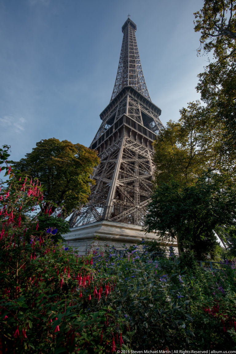 Oblique Eiffel Tower view behind colorful flowering plants and trees, with one iron leg and the upper tower visible.