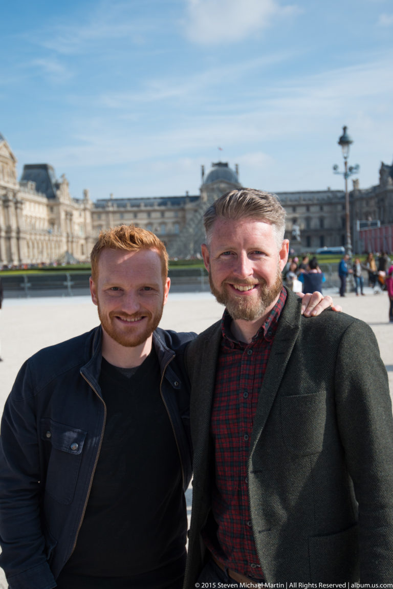 Two men posing together in the Louvre courtyard with the palace façades softly visible behind them under a blue sky.