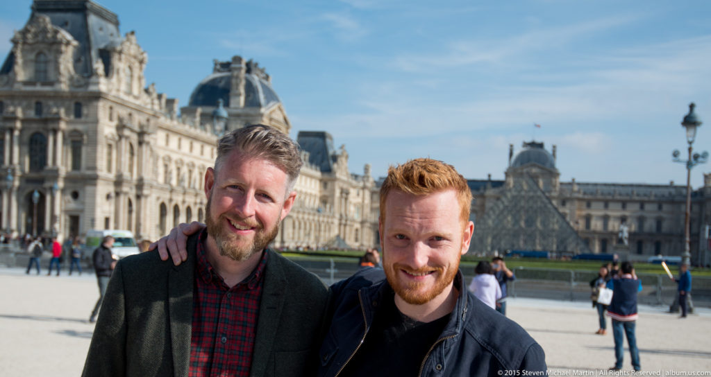 Two men posing in the Cour Napoléon with ornate Louvre Palace façades and rooflines extending across the background.