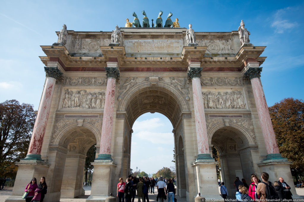 Frontal view of the Arc de Triomphe du Carrousel with three openings, pink columns, relief sculpture, rooftop statues, and visitors below.