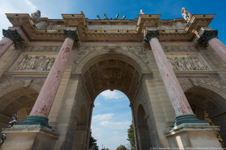 Low-angle view up through the Arc de Triomphe du Carrousel between pink marble columns, carved reliefs, and a blue-sky opening.