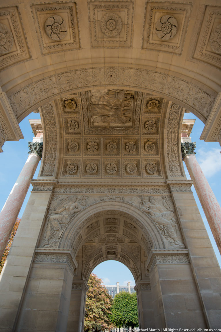 Oblique underside view of the Arc de Triomphe du Carrousel with coffered ceiling, pale stone arch, pink columns, trees, and sky.