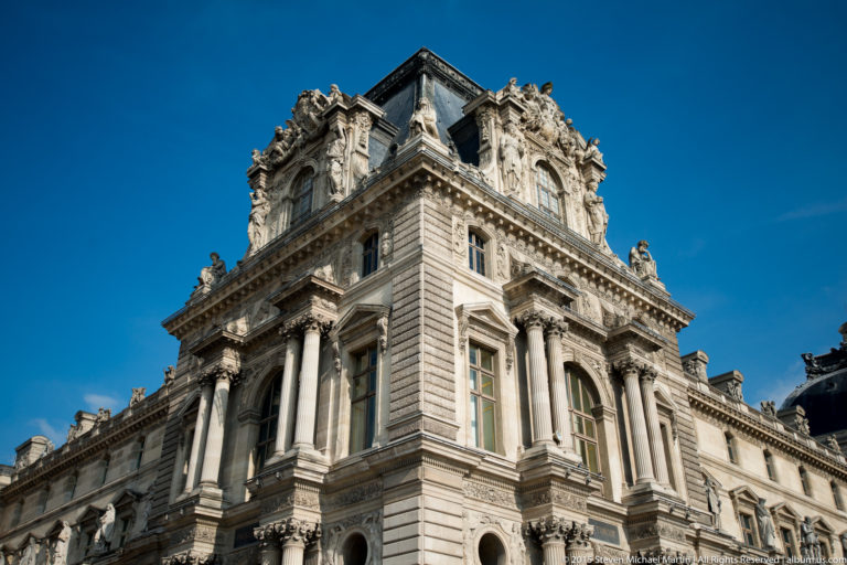 Low-angle view of an ornate Louvre Palace corner pavilion with columns, sculpture, rusticated stonework, and a steep roof against blue sky.