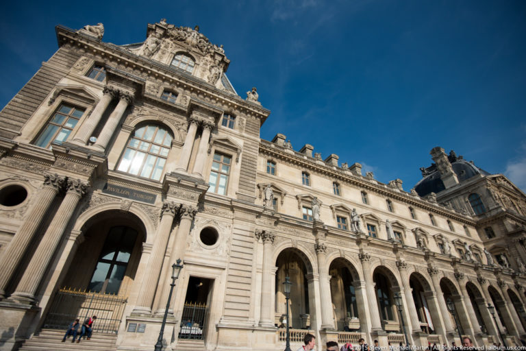 Wide low-angle view of the Louvre Palace façade with a monumental pavilion, arcaded wing, tall windows, columns, and rooftop sculpture.