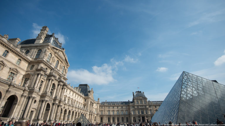 Louvre Pyramid in the Cour Napoléon beside ornate palace façades, with its glass-and-metal grid reflecting the blue sky.