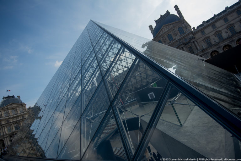 Tilted close view of the Louvre Pyramid with glass panels, dark metal framework, blue sky, and a palace roofline visible behind.