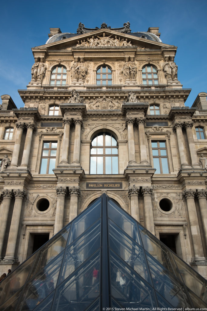 Pavillon Richelieu façade behind a pointed glass pyramid structure, with columns, round openings, sculpture, and the pavilion name visible.