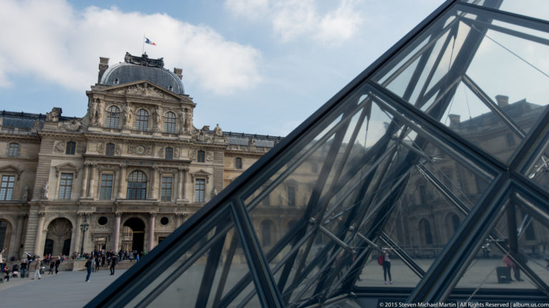 Diagonal glass face of the Louvre Pyramid crossing an ornate Louvre Palace façade, with metal grid lines, reflections, and blue sky.
