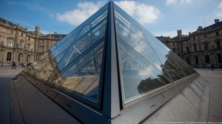 Small glass pyramid skylight in the Louvre courtyard with dark metal framing, cloud reflections, and palace façades around it.