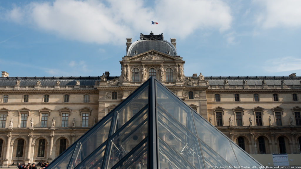 Small Louvre glass pyramid centered before a domed palace pavilion with the French flag, long stone façade, and cloudy blue sky.