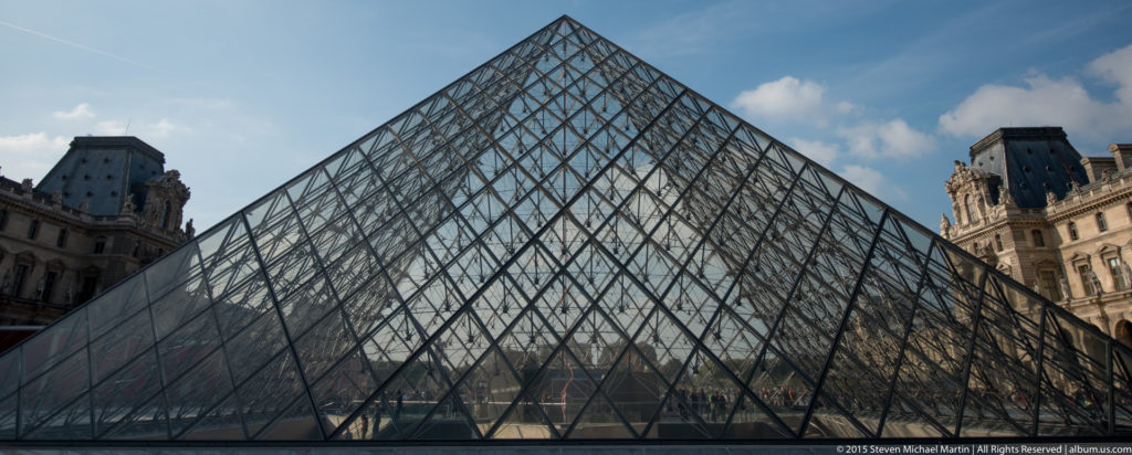 Close side view of the main Louvre Pyramid showing its large triangular glass surface, metal lattice, reflections, and palace edges.
