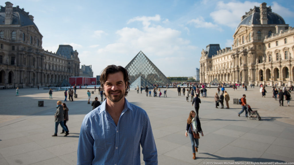 Man posing in the Louvre courtyard with the glass pyramid centered behind him and ornate palace wings extending across the background.
