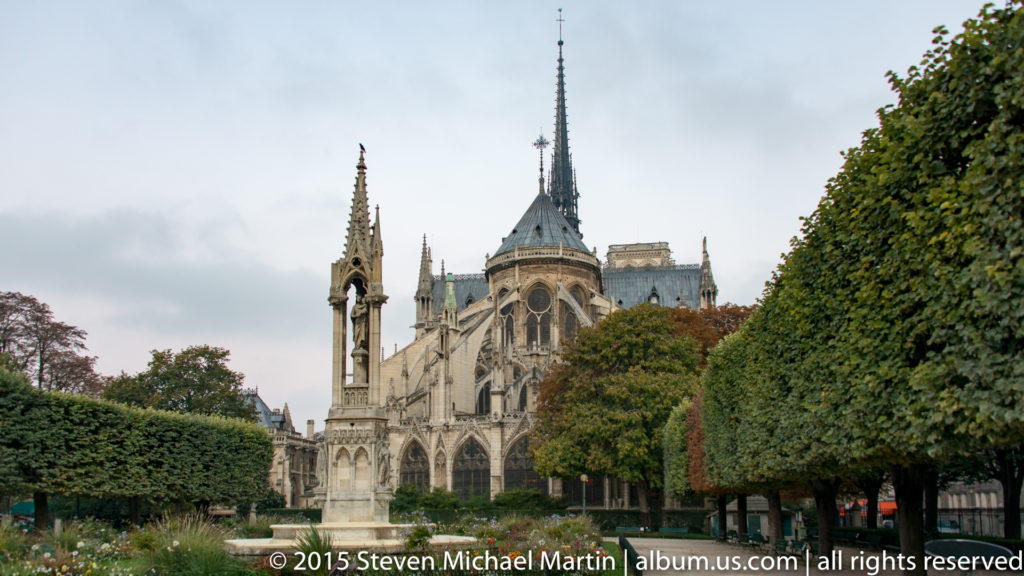 Distant garden view of the neo-Gothic Notre-Dame Fountain with the cathedral’s apse and spire beyond.