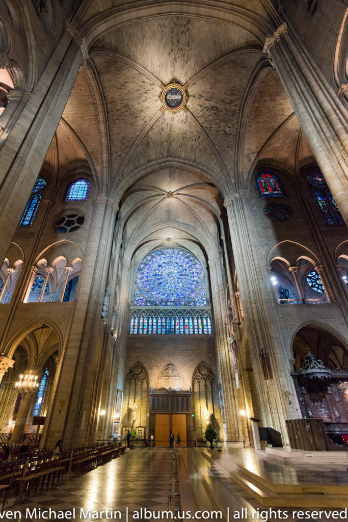 Wide view down the cathedral nave toward the west rose window, with rib vaults, piers, and side arcades.