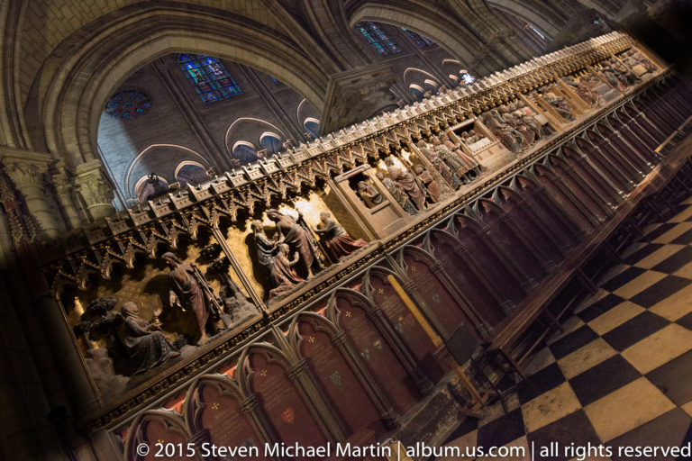 Diagonal view of a long sculpted choir enclosure above Gothic arcades, with checkered floor and cathedral interior beyond.