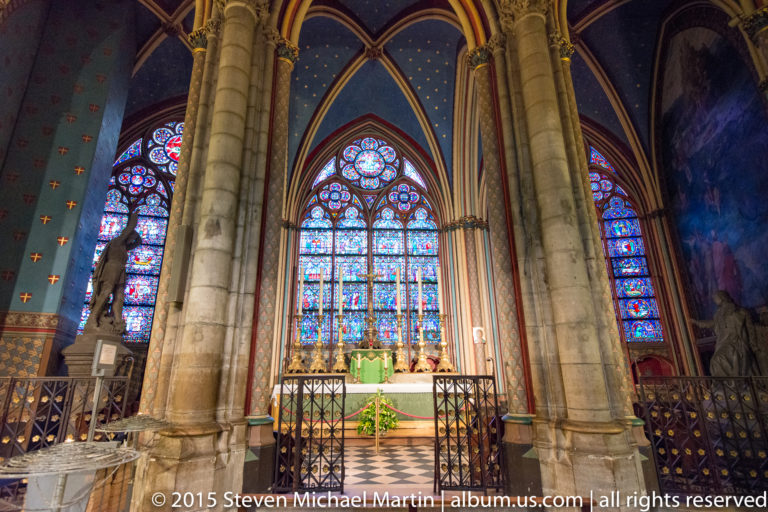 Gothic side chapel seen between large columns, with stained glass, altar furnishings, railings, and checkered floor.