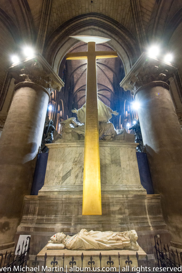 Tall luminous cross above the white-marble Pietà and choir sculpture at Notre-Dame de Paris.