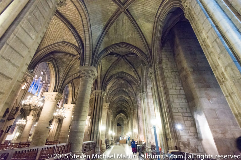 Long Gothic side aisle with pointed arches, rib vaults, clustered piers, chandeliers, and small visitors.