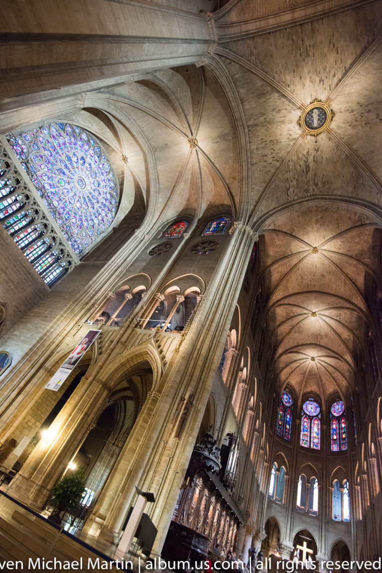 Diagonal low-angle interior with a rose window, tall piers, rib vaults, arcades, and distant stained glass.