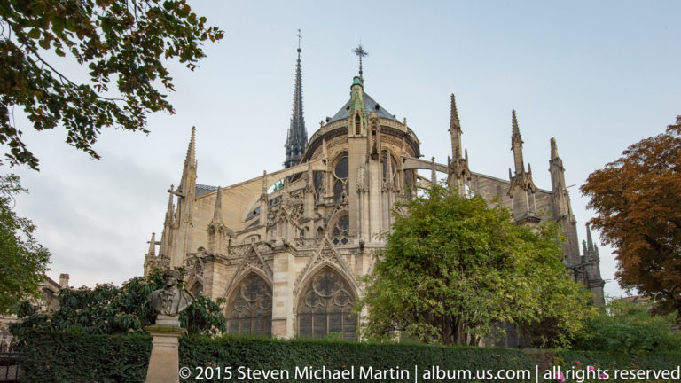 Rear three-quarter view of Notre-Dame with apse, flying buttresses, pinnacles, and central spire above trees.